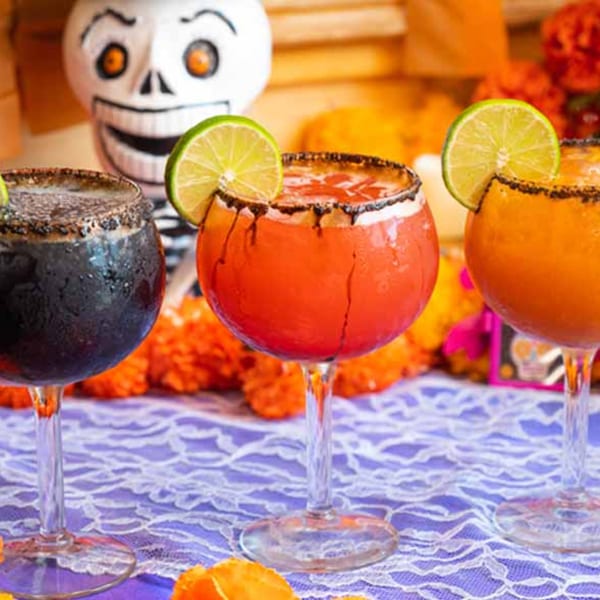 Three colorful margaritas with lime slices and chili rims, with a skeleton decoration in the background.
