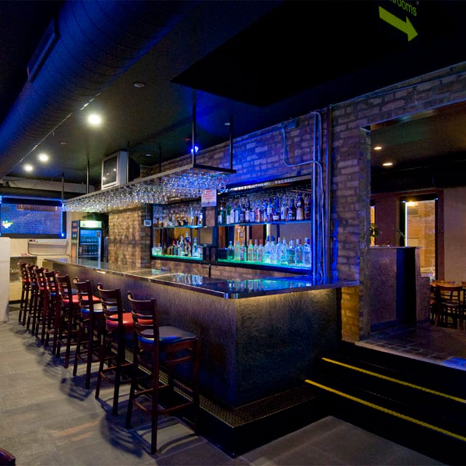 A long bar with stools, illuminated bottles, and blue lighting in Cesar's Killer Margaritas.