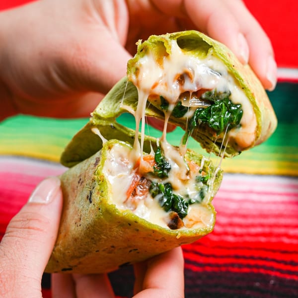 Hands pulling apart a green quesadilla, revealing melted cheese and spinach, against a colorful striped background.