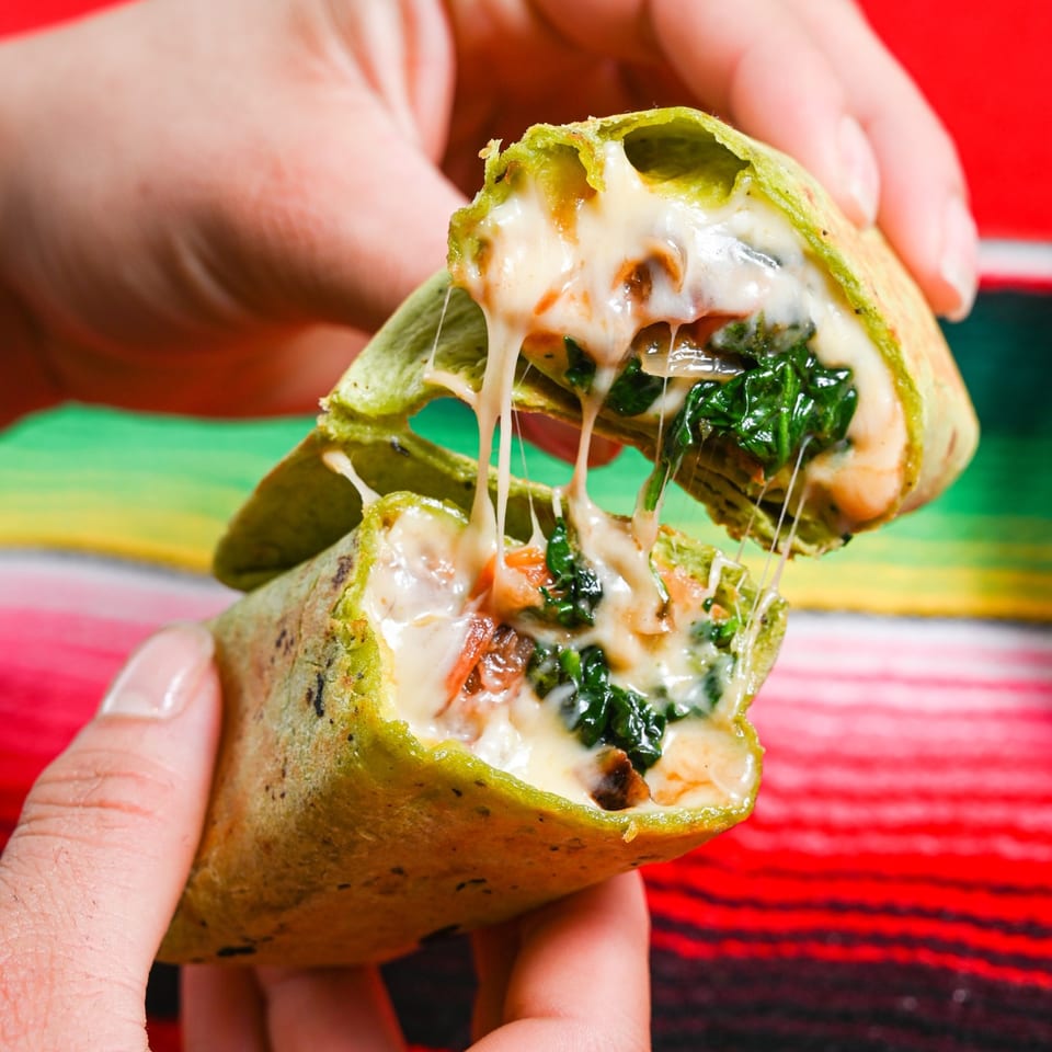 Hands pulling apart a green quesadilla, revealing melted cheese and spinach, against a colorful striped background.