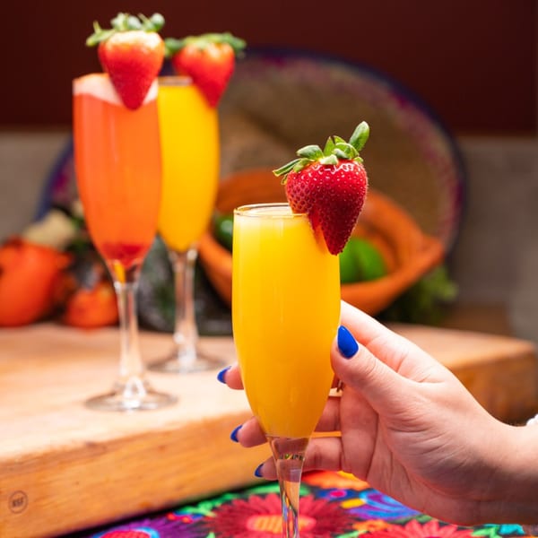 A hand with blue nails holds a mimosa garnished with a strawberry, with two more drinks behind it.