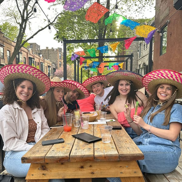 Six smiling people in sombreros sit at a wooden picnic table on an outdoor patio decorated with colorful papel picado.
