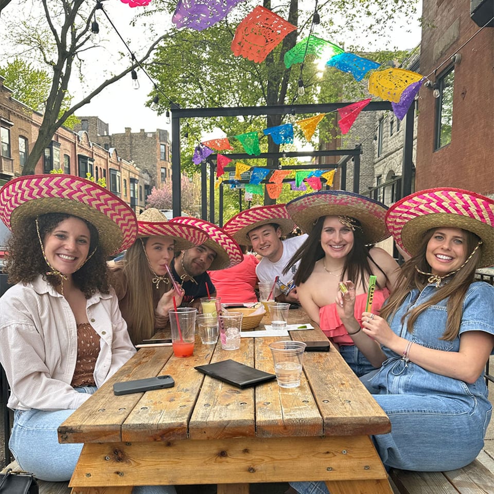 Six smiling people in sombreros sit at a wooden picnic table on an outdoor patio decorated with colorful papel picado.