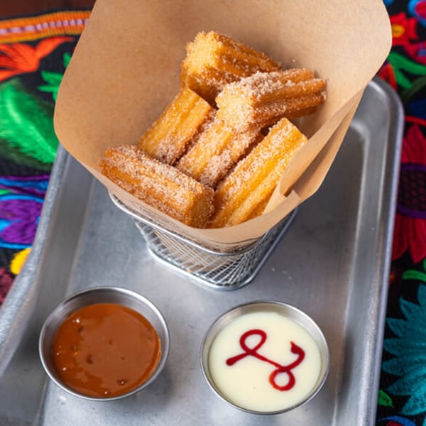Cinnamon sugar churros in a metal basket with caramel and cream dipping sauces on a tray.
