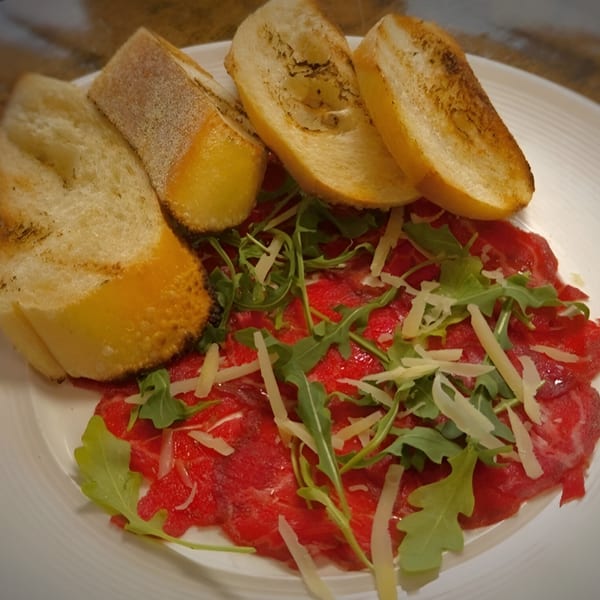 Thinly sliced red meat carpaccio with arugula, parmesan cheese, and toasted bread on a white plate.