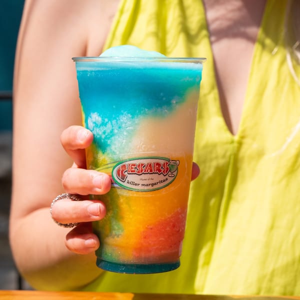 A hand holds a vibrant, multi-colored frozen margarita from Cesar's Killer Margaritas.