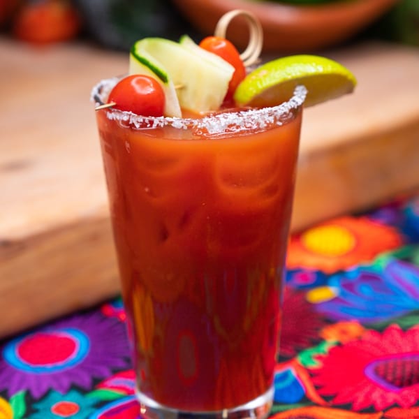 A Bloody Mary cocktail garnished with cherry tomatoes, cucumber, and lime, on a colorful floral tablecloth.