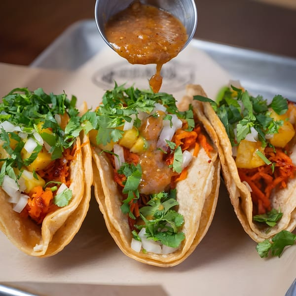 Three Mexican tacos with pineapple, onions, cilantro, and sauce being poured from a metal cup.