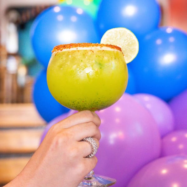 Hand holding a green margarita with a chili-lime rim and lime slice, against blue and purple balloons.