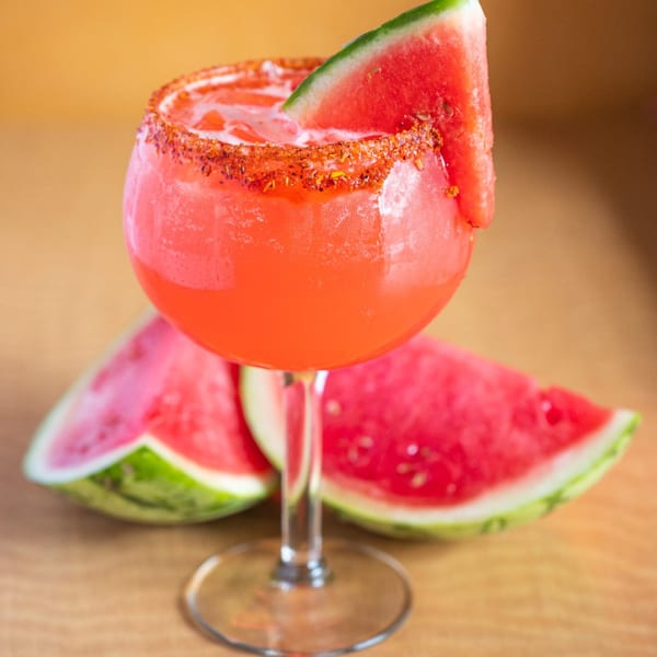 Watermelon margarita with a chili-lime rim and a watermelon slice, with watermelon wedges in the background.