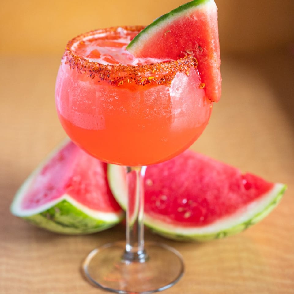 Watermelon margarita with a chili-lime rim and a watermelon slice, with watermelon wedges in the background.