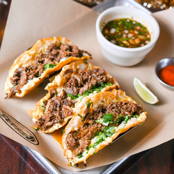 Three birria tacos with cilantro and onions, served with consommé, hot sauce, and a lime wedge.