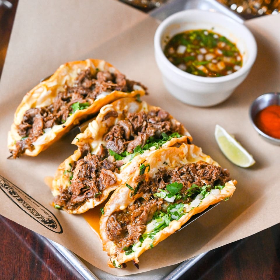 Three birria tacos with cilantro and onions, served with consommé, hot sauce, and a lime wedge.