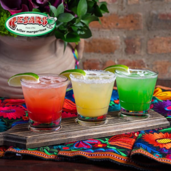 Three colorful margaritas, red, yellow, and green, with lime wedges on a wooden tray and vibrant tablecloth.