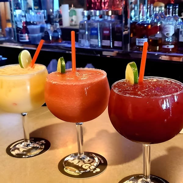 Three colorful margaritas, yellow, orange, and red, with lime garnishes and straws on a bar.