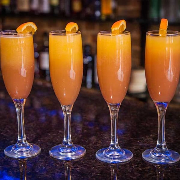 Four champagne flutes filled with orange and red layered drinks, garnished with orange slices, on a bar.