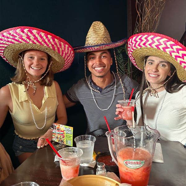 Three smiling customers wearing sombreros and pearl necklaces, enjoying margaritas at Cesar's.