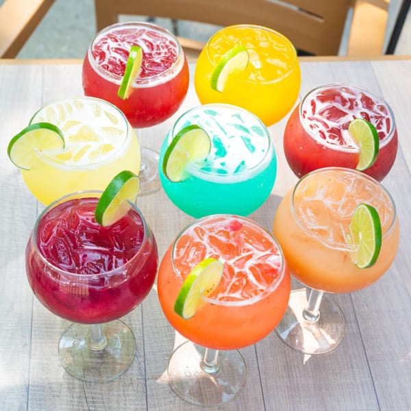 Nine colorful margaritas with lime garnishes, including red, yellow, blue, and orange, on a light wooden table.