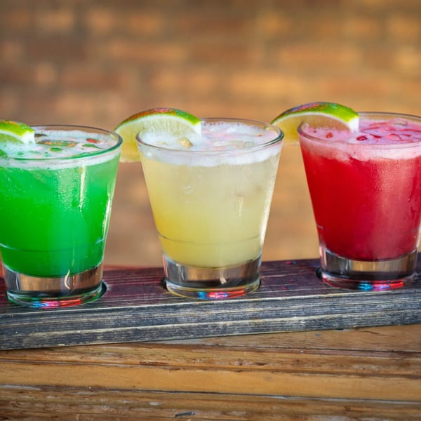 Three colorful margaritas, green, yellow, and red, garnished with lime wedges on a wooden tray.